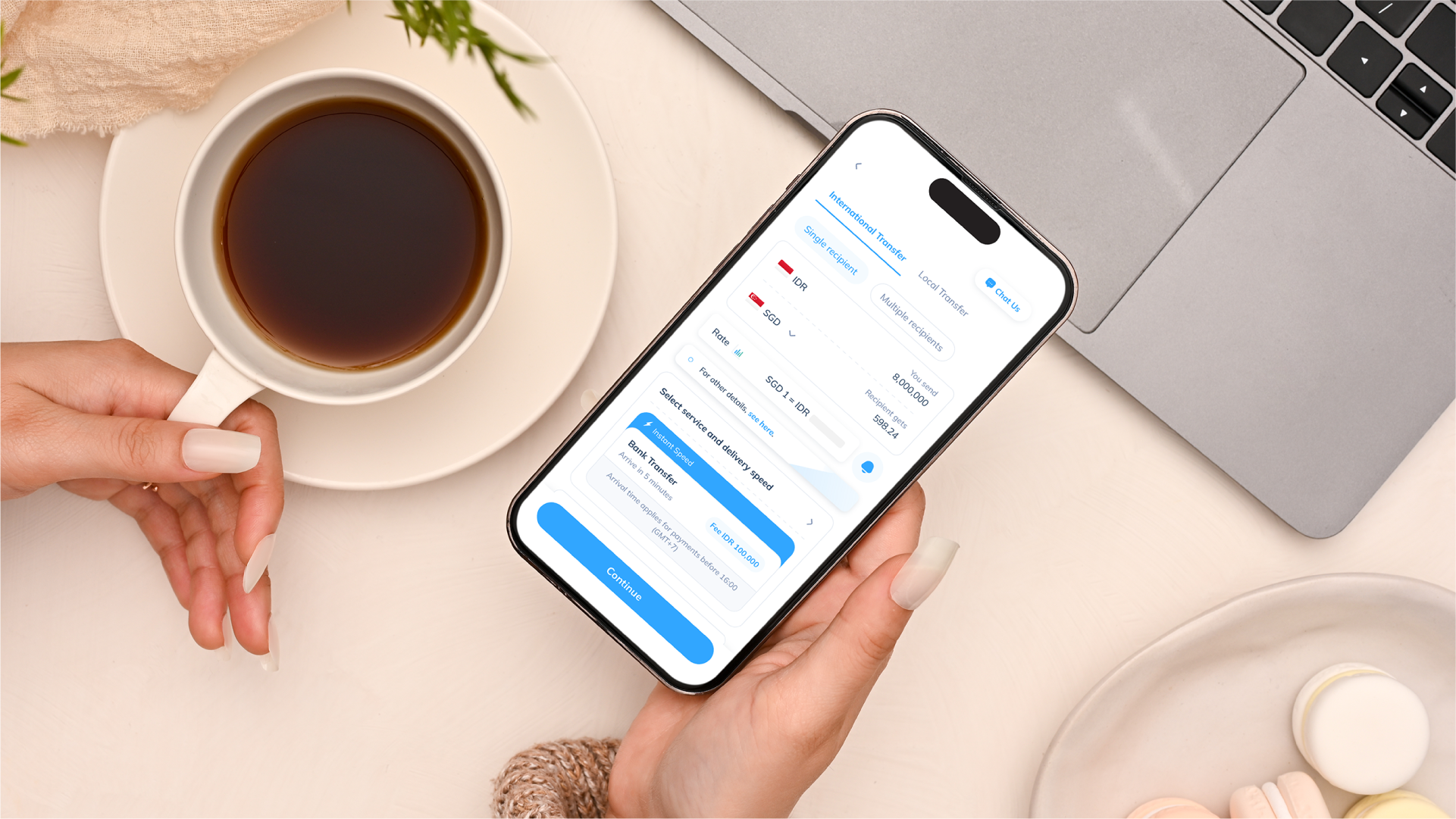 Seseorang memegang smartphone yang menampilkan aplikasi transfer internasional di atas meja dengan secangkir kopi, laptop, dan sepiring macaron.