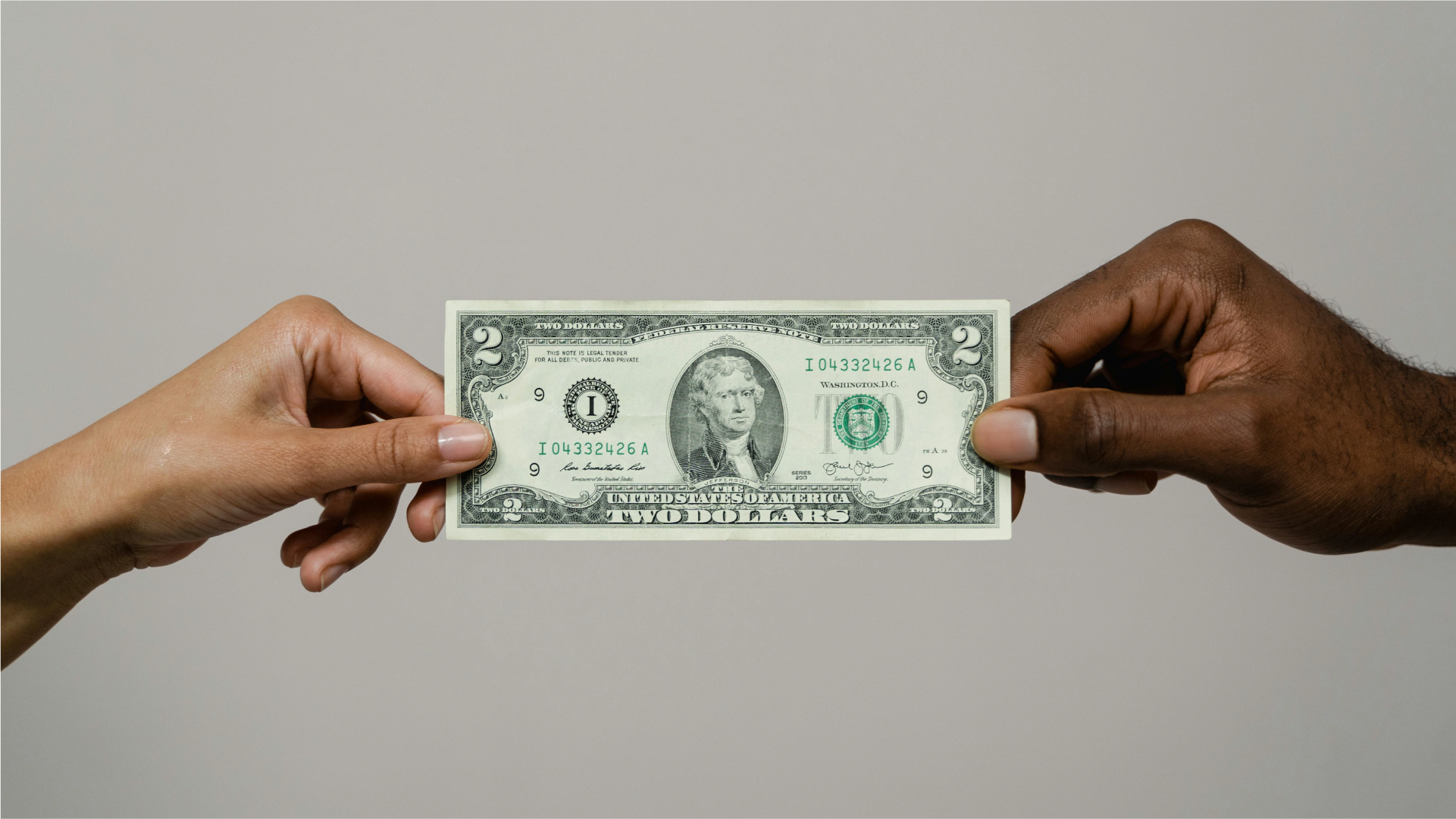 Dua tangan memegang sisi kiri dan kanan selembar uang 2 dolar Amerika Serikat melambangkan transaksi keuangan.