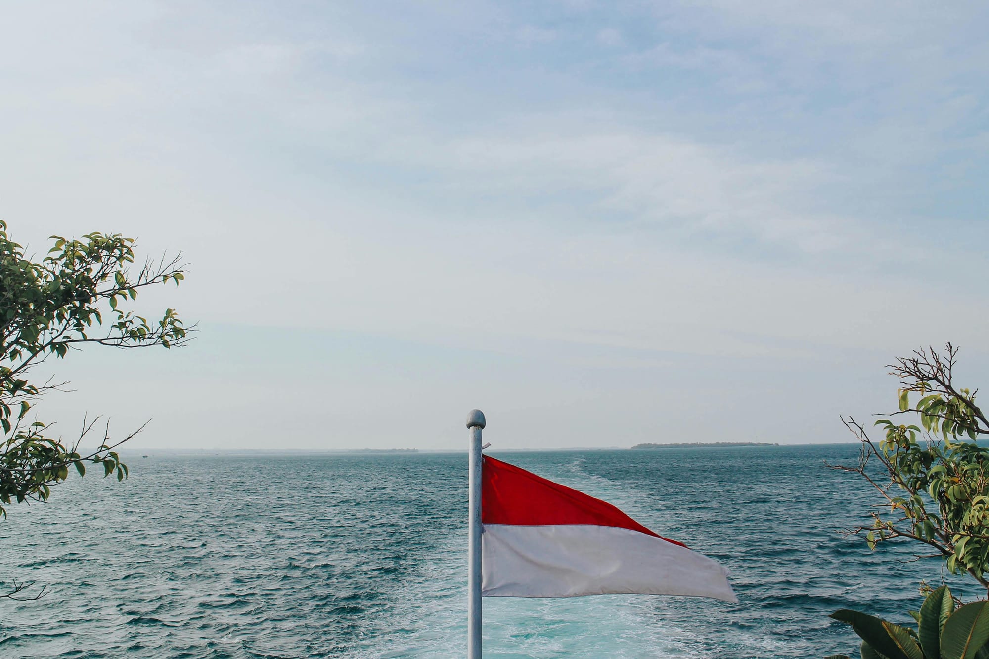 Red and white Indonesian flag standing by the ocean under a bright sky.