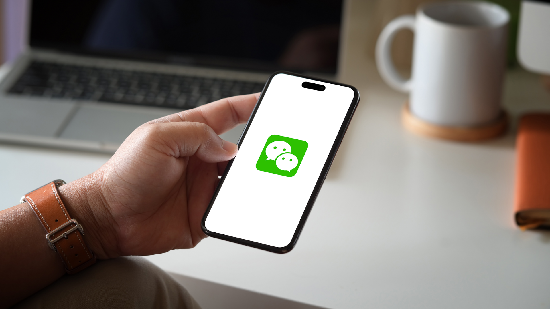 Tangan memegang smartphone dengan logo WeChat di dekat laptop sebagai ilustrasi akses WeChat Pay melalui ponsel.