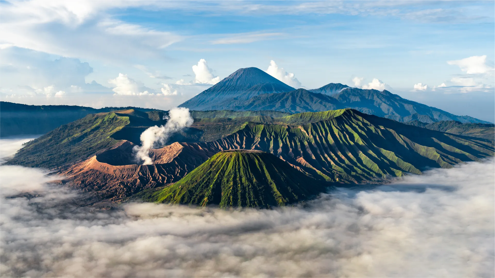 Gunung Bromo 的火山景观,火山口冒着白烟,周围是绿色山丘和 Bromo Tengger Semeru 地区的云海。