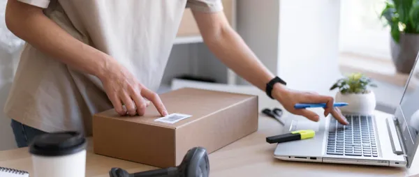 Potret seseorang yang sedang memegang pulpen sambil memeriksa paket yang dipesan pembeli secara online; di depannya ada lapt 